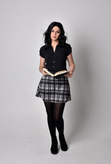 Portrait of a goth girl with dark hair wearing blue and plaid skirt with boots. Full length standing pose, holding a book, on a studio background.