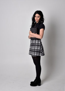 Portrait of a goth girl with dark hair wearing blue and plaid skirt with boots. Full length standing pose, holding a book, on a studio background.