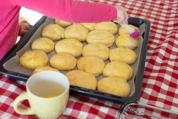 Child girl in a kitchen glazing bread buns with egg wash before baking. Lockdown activity idea for preschoolers.