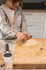 Fototapeta na wymiar Child girl, 3 years old, in white kitchen, flattening pizza dough with a rolling pin on a wooden board. Lockdown activity idea for preschoolers.