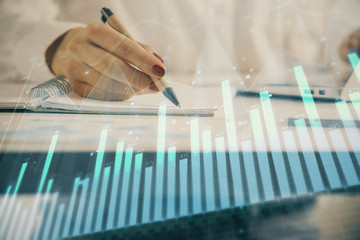 Double exposure of woman's hands making notes with forex graph hologram. Concept technical analysis.