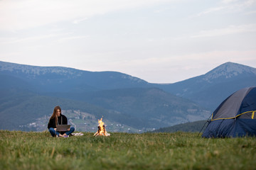 Fototapeta premium young female freelancer working on laptop in the mountains in the evening. Tourist girl sitting near campfire and having fun. Copy space.