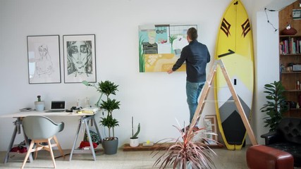 Hipster man at home alone, hangs painting on white wall. Trendy and modern loft apartment, beautiful and authentic interior design. Remodelling or support local artists, cosy vibes