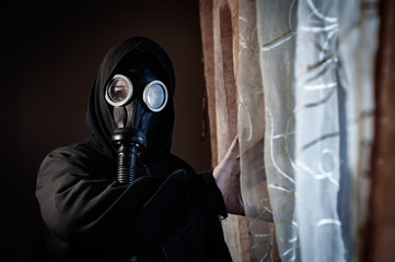 Unknown man wearing black hood and protective gas mask standing by the window at home looking to...