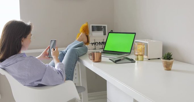 Businesswoman relaxing and using phone after finished work. Put feet up on the desk in home office. MacBook Pro Laptop with Green Chroma Key Screen Stands on a Desk in the Living Room. Cozy apartment