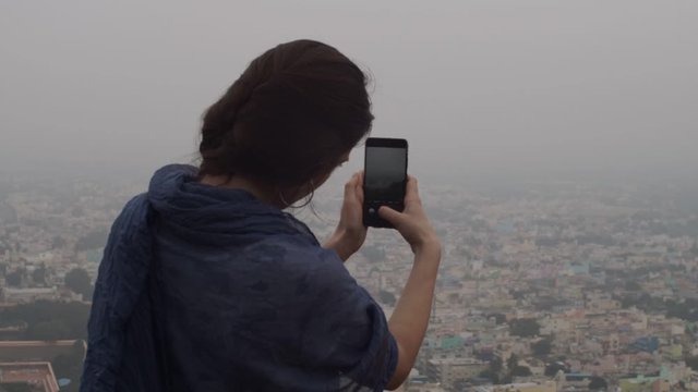 Back view of young woman making photo of misty cityscape on telephone outdoors copy text space. Unrecognizable brunette girl holding mobile phone shooting panoramic city slow motion. Social media