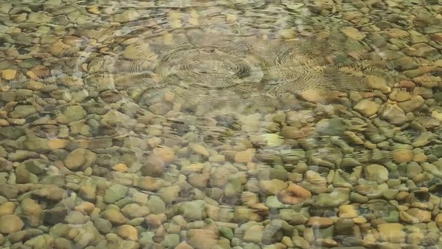 Water drop on crystal clear pebbles beach pond with beautiful ripples, top view. Abstract nature background of rock stone lake bottom with transparent water & slowly raindrops, Close up - Slow motion 