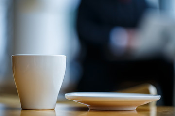A white cup and saucer with hot aromatic coffee stands on a wooden table against the background of a sitting businessman