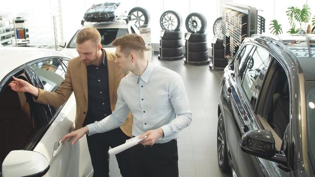 Young affable consultant talking about features of new car to customer. Business man listen to him, choosing new auto in dealership.