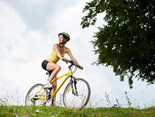 Attractive happy woman cyclist riding on yellow mountain bicycle on a grassy hill, wearing helmet,...