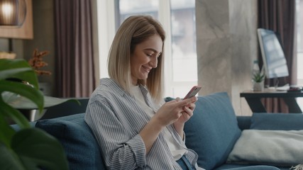 Caucasian young woman sitting on couch in modern home using phone and smiling texting message watching contents or bloggs scrolling tapping apartment technology apps shopping sunlight sunny day