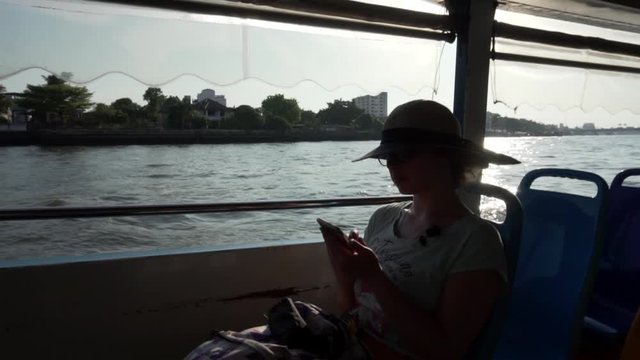 A young girl in a hat sits at the edge of the side of a pleasure boat and looks into the distance. Silhouetted girl sitting in the cabin of the ferry, on the background of river water