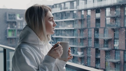Close up view of cheerful young woman in white hoodie stays in balcony drink hot coffee or tea relax enjoy breathing fresh air healthy freezy snow falls in march morning no stress concept