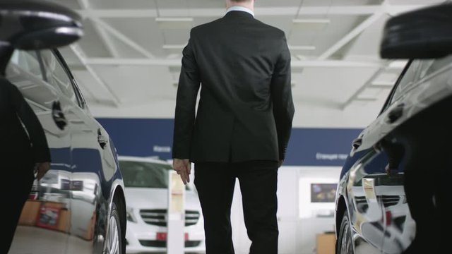 Young handsome auto dealer posing in car dealership. He is walking between new cars and then turns around looking at camera and smiling.