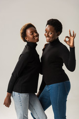smiling african american girls in black turtlenecks showing ok sign isolated on grey