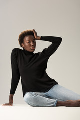 african american girl in black turtleneck sitting on white cube isolated on grey