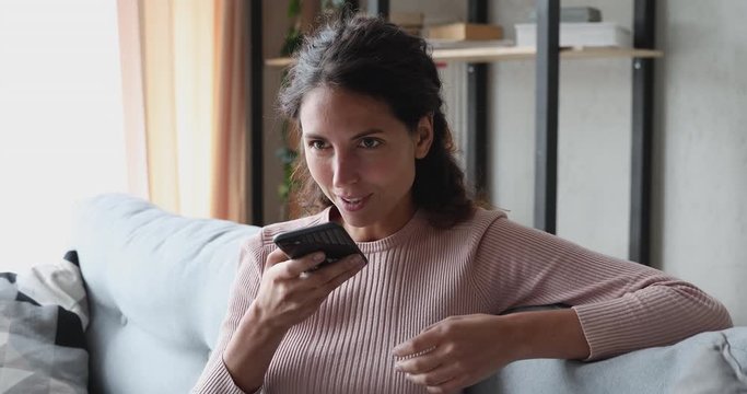 Smiling young woman using voice recognition assistant on smartphone at home. Happy lady records audio message, talking on mobile speakerphone, commands digital ai assistance app online search concept.