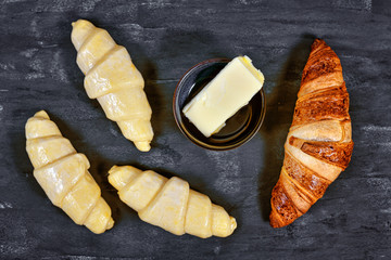 Freshly baked croissant pastry next to unbaked cresent shaped dough layered with butter on dark background