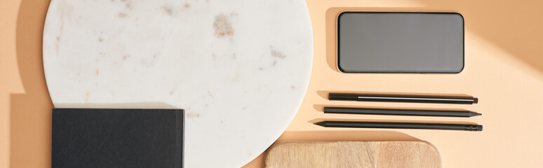panoramic shot of wooden and marble boards, smartphone, notebook and pens on beige background
