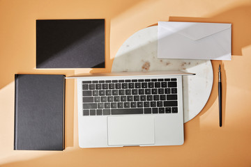 top view of laptop on marble board, black notebooks, envelope and paintbrush on beige background