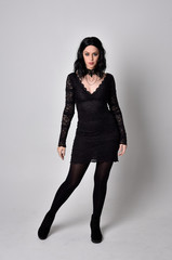 Portrait of a goth girl with dark hair wearing black lace dress and boots. Full length standing...