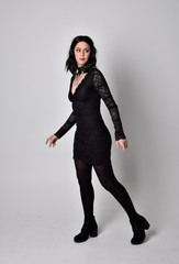 Portrait of a goth girl with dark hair wearing black lace dress and boots. Full length standing pose on a studio background.