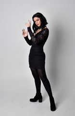 Portrait of a goth girl with dark hair wearing black lace dress and boots. Full length standing...