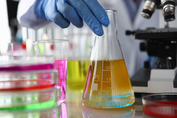 Close-up of chemist hand holding flask with yellow liquid. Researcher making analysing of liquor sample in laboratory. Chemistry industry and science test concept