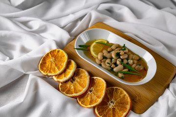 Marinated white beans in their own juice on an oval plate with Thai pineapple sauce and green onions. Decorated with a slice of lemon and dried oranges on a wooden Board with a snow white fabric