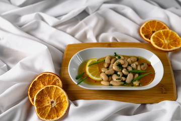 Marinated white beans in their own juice on an oval plate with Thai pineapple sauce and green onions. Decorated with a slice of lemon and dried oranges on a wooden Board with a snow white fabric