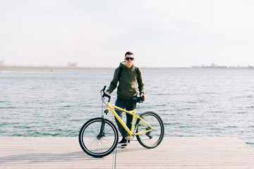 a young man in khaki sportswear and glasses stands on a pontoon near the lake and holds a yellow Bicycle near him on a Sunny spring day