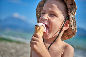 White blond boy with hat at seaside eating or licking ice cream with shallow depth of field and...
