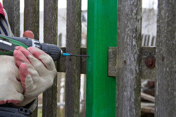 a man spins a screw into a wooden board with a screw. The green-painted board is screwed into an old fence