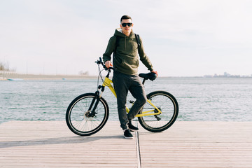 Obraz premium a young man in khaki sportswear and glasses stands on a pontoon near the lake and holds a yellow Bicycle near him on a Sunny spring day