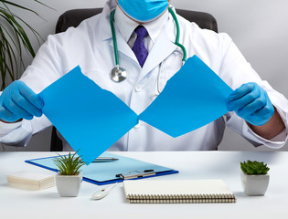 doctor in a white medical coat is sitting at a table in a brown leather chair and holding a torn blue sheet of paper in his hands