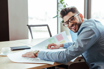 UI designer smiling and looking at camera with wove papers near cup of coffee and notebook at table