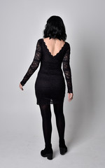 Portrait of a goth girl with dark hair wearing black lace dress and boots. Full length standing pose, facing away from the camera, on a studio background.