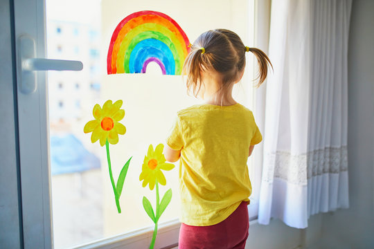 Adorable toddler girl attaching drawing of rainbow to window glass as sign of hope