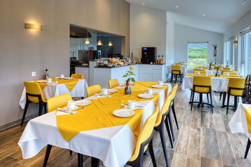 Elegant and minimalist restaurant in a Scandinavian hotel with fantastic views of the countryside and mountains. Yellow and white tablecloths with breakfast cutlery. Large windows.