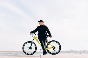 a young man in black sportswear stands on a pontoon near the lake and holds a yellow Bicycle near him on a Sunny spring day