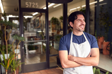 Smiling Male Owner Of Florists Standing In Doorway Surrounded By Plants