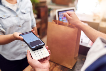 Close Up Of Woman In Store Making Contactless Payment At Sales Desk Holding Mobile Phone To Reader