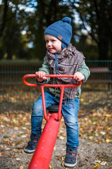 Little cheerful boy in a blue cap on a red seesaw