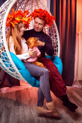 Beautiful brunette girl whth long hair in the blue shirt and man in black sweater with a wreaths of yellow autumn maple leaves and small dog on a white wicker hanging chair with pillow