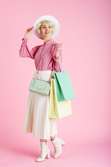 Spring shopping concept: happy smiling fashionable woman wearing trendy clothes posing with colorful paper bags. Full-length portrait. Pastel pink color background. Copy, empty space for text