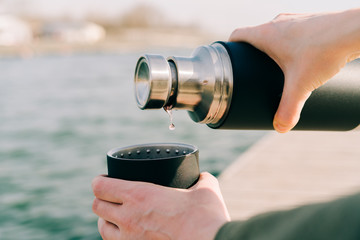 the guy is holding a black stylish eco-friendly stainless steel thermos and pours hot, herbal tea into a mug outdoors on a Sunny spring day near the lake