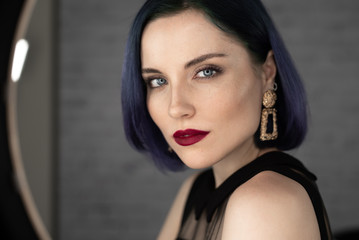 Fototapeta na wymiar Young beautiful girl with professional makeup, blue hair and perfect skin sits in studio in front of ring light lamp posing for social media. Beauty blog. Copy space.