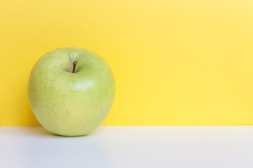 Single fresh green apple isolated on yellow background and white table. Free space for text.