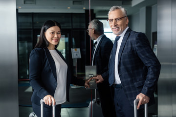 smiling businessman and asian businesswoman with travel bags pushing button of elevator