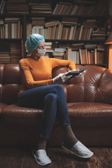 Woman with protective antiviral mask sitting at home in isolation / quarantine and reading book.